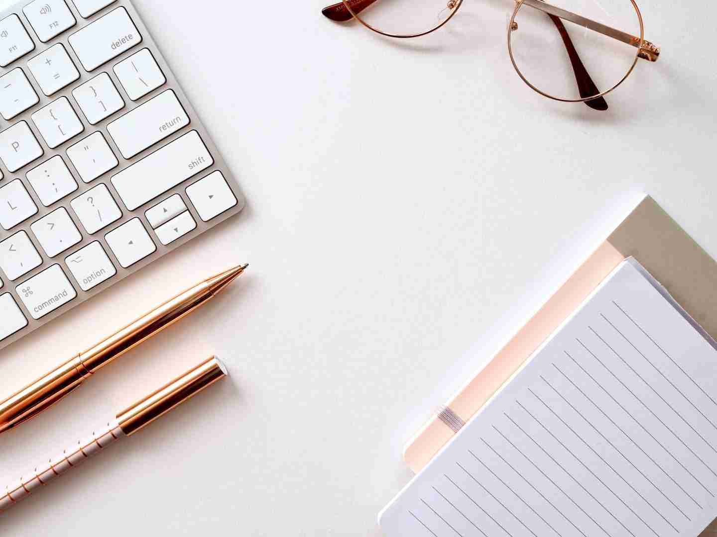 A desk with writing utilities, including two pens, two notebooks, a laptop and a pair of glasses.