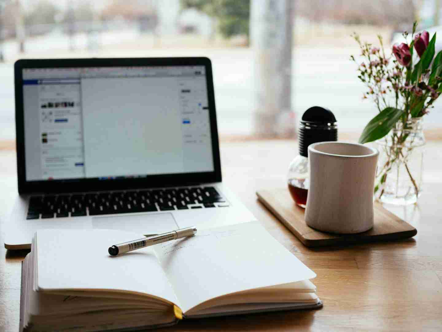 A desk with a laptop, a plant, a cup of drink and a notebook on top of it. There is a pen on top of the notebook.