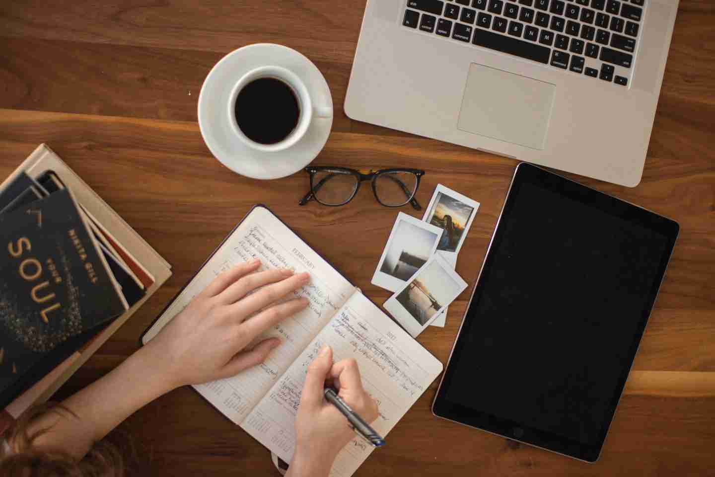 A desk filled with a laptop, a stack of books, a pair of glasses, three polaroids, an iPad, a cup of coffee and a notebook. There is someone writing in the notebook.