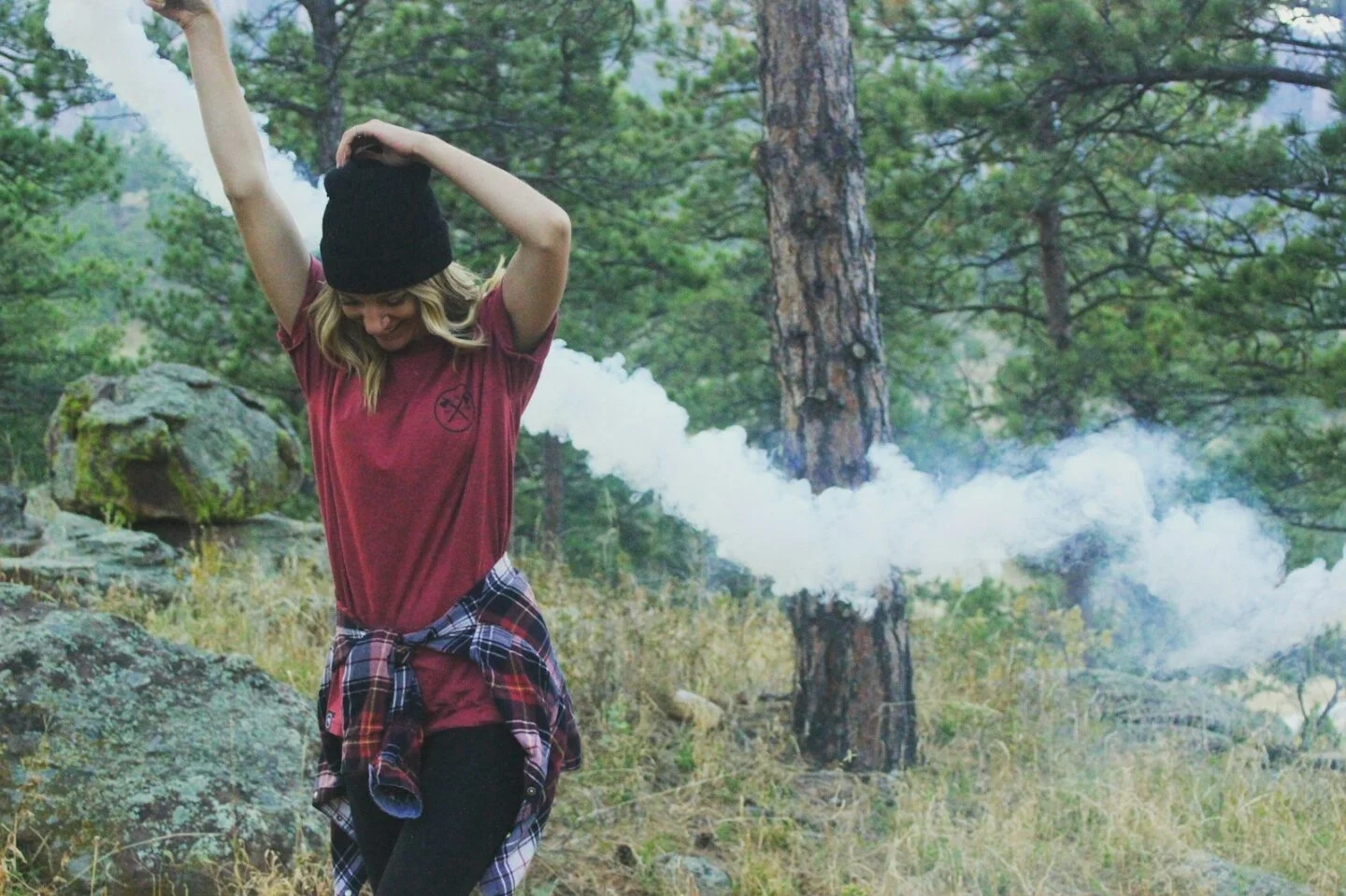 woman raising arms holding white smoke bomb standing beside rock surrounded with trees at daytime